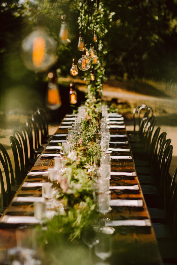 Outdoor wood wedding reception table with black chairs and edison bulb lights above it